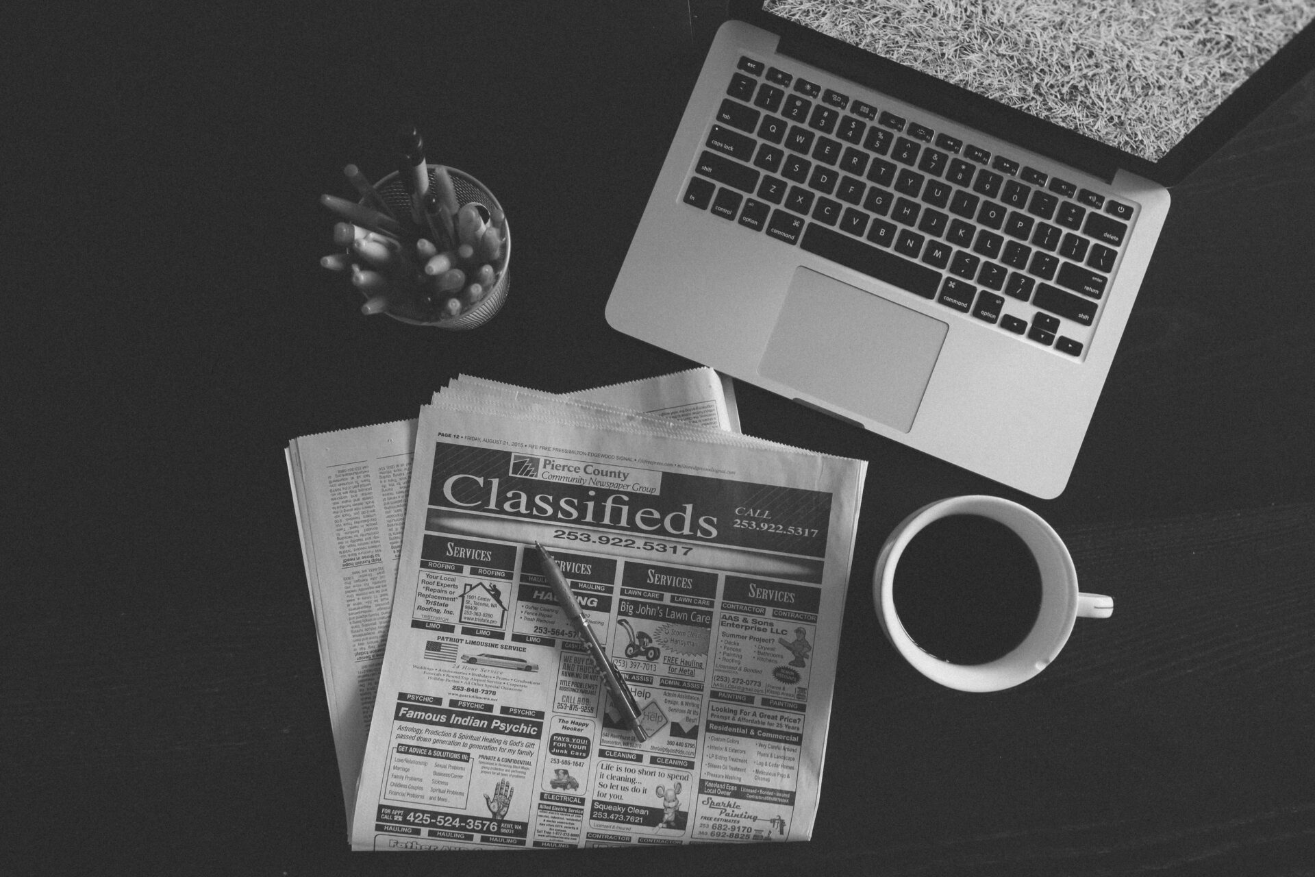 A black and white image of a laptop, newspaper, and coffee cup on a desk.